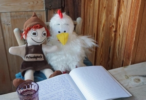 die kleine Handpuppe Schokopuck sitzt zusammen mit der Handpuppe Eggy Bird, ein weißes Huhn, am Tisch vor einem handgeschriebenen Text auf dem Balkon
